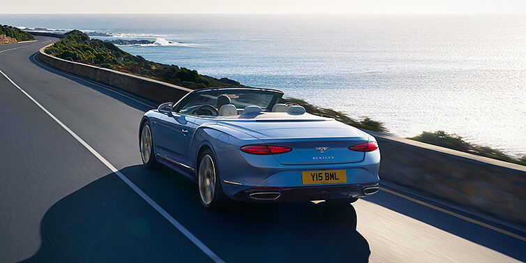 Bentley Copenhagen New Bentley Continental GTC Azure convertible rear view in Light Windsor blue paint with 22 inch ten spoke wheels driving dynamically on an ocean road in the sunshine
