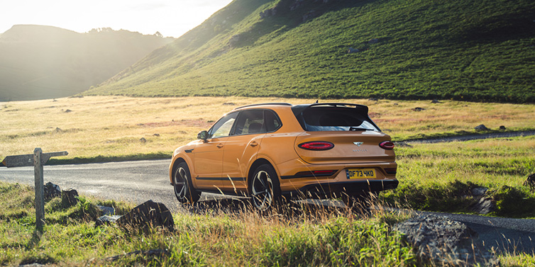 Bentley Copenhagen Bentley Bentayga S SUV rear three quarter in Sunburst Gold paint on a road surrounded by green hills