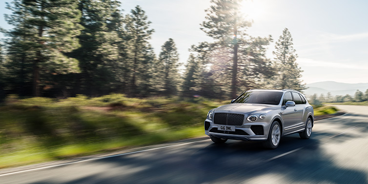 Bentley Copenhagen Bentley Bentayga SUV front three quarter in Moonbeam silver paint driving dynamically along a tree-lined road