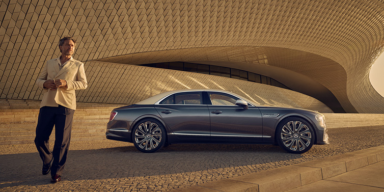 Bentley Copenhagen Bentley Flying Spur Mulliner sedan in Rose Gold over Magnetic duotone paint parked by a curved wall with person in suit jacket standing on the left